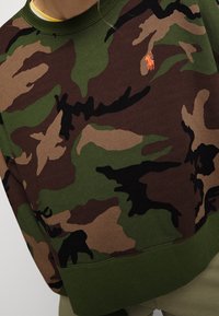 Camouflage sweatshirt in green, brown, and black with ribbed hem and collar. Features an orange embroidered logo on the front.