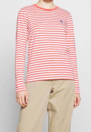 Person wearing a long-sleeve coral and white horizontal striped shirt with beige pants, standing against a plain light background.