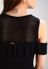 Top nero con design a rete sulla schiena, dettagli arricciati lungo le spalle e un logo metallico vicino alla vita.