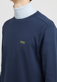 Námořnická mikina s žebrovým výstřihem, na které se nachází žluté logo "BOSS". Měkká textura, volný design, přiléhavý silueta a strukturované ramenní švy.