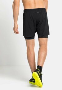 Zwarte sportshorts met een aansluitende binnenlaag, elastische tailleband en zichtbare stiksels. Draagt zwarte en neon gele hardloopschoenen.