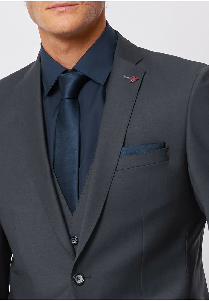 Veste de costume gris foncé avec une texture lisse, associée à une chemise bleu marine et une cravate. Le mouchoir de poche s'accorde avec la cravate. Détail en métal argenté sur le revers.