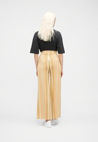 Άτομο με μακριά ξανθά μαλλιά στέκεται με την πλάτη, φορώντας μαύρο κοντομάνικο τοπ, φαρδιά παντελόνια με ρίγες σε μπεζ και πορτοκαλί χρώμα και λευκά αθλητικά παπούτσια.