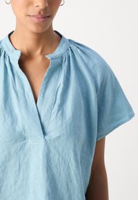 Femme portant un chemisier bleu clair à manches courtes avec une encolure fendue et de petites fronces près du col.