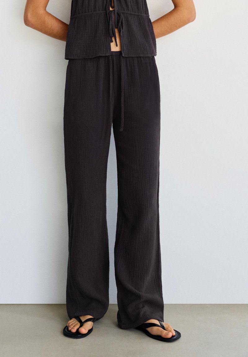Femme portant un pantalon noir ample texturé et des sandales noires en tongs, debout contre un mur blanc, les bras dans le dos.