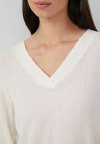Vrouw in een gemêleerde witte V-hals top, met een zichtbare sleutelbeen en nek, met steil donkerbruin haar.