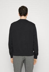 Sudadera negra de algodón, con cuello redondo y puños de canalé, vista desde la parte posterior, combinada con pantalones de chándal grises.