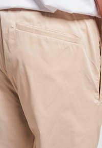 Pantaloni din bumbac bej, cu o textură fină, având un buzunar pe spate și o croială lejeră. Materialul are un ușor luciu.