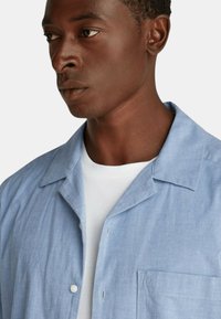 Camicia azzurra a maniche corte con colletto a punta, bottoni frontali e taschino sul petto. Il tessuto ha una consistenza liscia con dettagli di cucitura minimi.