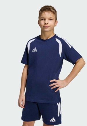 Ragazzo giovane in piedi con la mano sui fianchi, indossa abbigliamento sportivo Adidas blu navy e bianco, inclusa una t-shirt e pantaloncini.