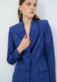 Blazer texturé bleu avec un motif floral en relief, larges revers, trois boutons et une coupe ajustée, comprenant des poches à l'avant.