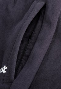 Gros plan d'une poche de sweat à capuche sombre avec une fermeture éclair noire partiellement ouverte, montrant la texture douce et tricotée du tissu ainsi que le bord du texte brodé en blanc.