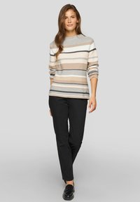 Frau geht selbstbewusst in einem beigefarbenen, weißen und grauen gestreiften Pullover, schwarzen Hosen und schwarzen Ballerinas vor einem hellgrauen Hintergrund.