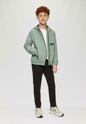 Veste à capuche vert clair avec fermeture éclair noire et poche, t-shirt blanc, pantalon noir et baskets beiges, le tout sur un fond uni.