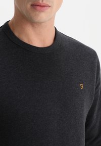 Maglione grigio scuro a girocollo realizzato in tessuto morbido, con un piccolo logo arancione sul lato sinistro del petto. Texture senza cuciture.
