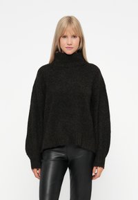 Übergrößter schwarzer Strickpullover mit hohem Rollkragen, sanfter Textur und weiten Ärmeln, kombiniert mit schwarzen Lederhosen.