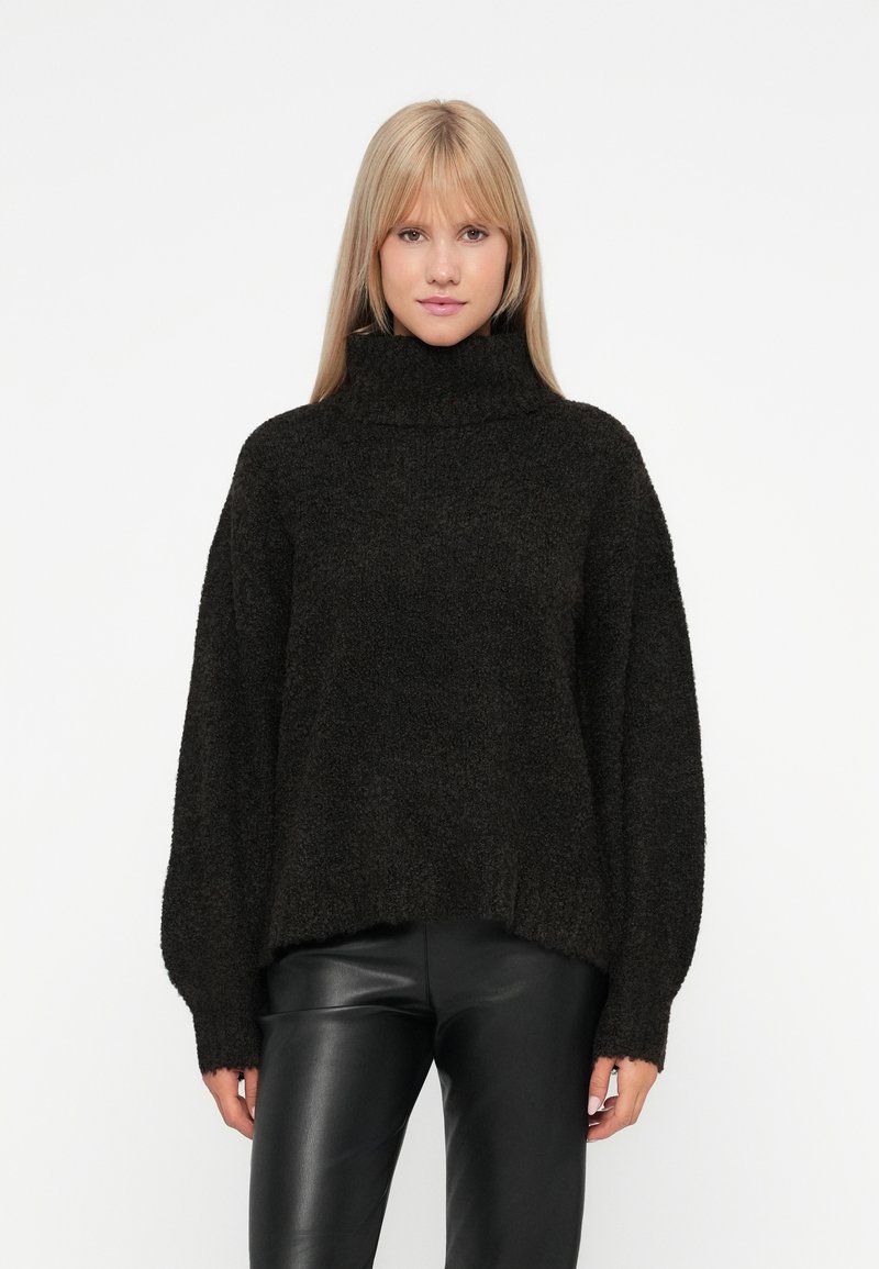 Übergrößter schwarzer Strickpullover mit hohem Rollkragen, sanfter Textur und weiten Ärmeln, kombiniert mit schwarzen Lederhosen.