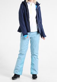 Jacke mit Marine-Muster und Reißverschluss, weißes Oberteil und hellblaue Schneehosen. Schwarze Sneakers runden das Outfit ab.