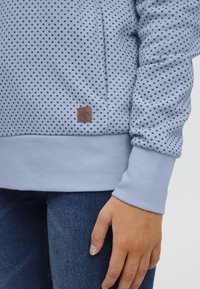 Felpa azzurra con un motivo a pois, dotata di polsini e orlo inferiore a costine, e una patch in pelle marrone con il logo sul lato.