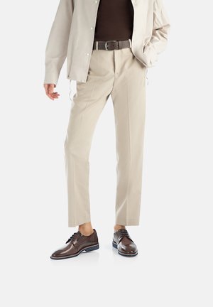 Pantaloni beige con una vestibilità dritta e piega, abbinati a scarpe in pelle marrone scuro con una finitura liscia e accenti minimi.