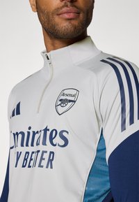 Hvid atletisk halv-zip trøje med blå accenter, Arsenal-logo og teksten "Emirates Fly Better". Glat stof med subtil tekstur og striber.
