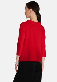 Top rosso a maniche tre quarti con scollo rotondo. Tessuto morbido, vestibilità rilassata e design semplice. Indossato con pantaloni scuri a trama.