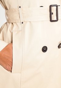 Main dans la poche d'un trench-coat beige avec ceinture et boutons foncés, montrant des coutures détaillées et la texture du tissu.