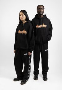 Sudaderas con cremallera negra y gráfico de llamas y pantalones de chándal negros con logotipos. Ambos tienen un ajuste holgado y un diseño minimalista, combinados con calzado negro.