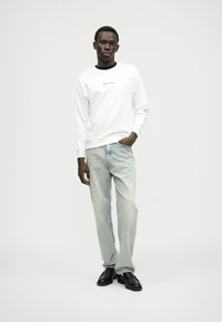 Balts džemperis ar melnu apkakli un logo, kombinēts ar gaiši ziliem džinsiem un melnām kurpēm. Audums izskatās mīksts un ir brīvs fit.