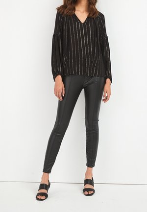 Femme portant un chemisier rayé noir à manches longues, un pantalon en cuir noir et des sandales noires à bouts ouverts, debout devant un fond blanc.