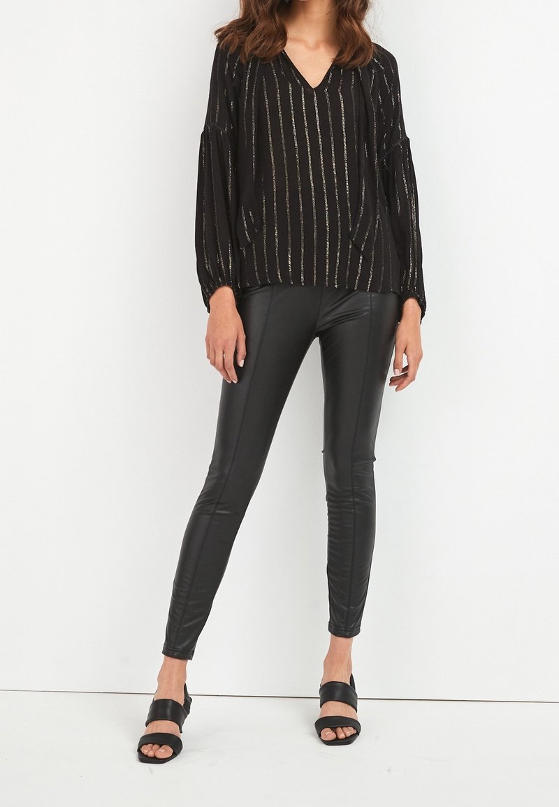 Femme portant un chemisier rayé noir à manches longues, un pantalon en cuir noir et des sandales noires à bouts ouverts, debout devant un fond blanc.
