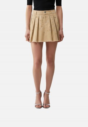 Donna che indossa una minigonna plissettata beige con ricami floreali, un top nero e sandali argentati con tacco alto e cinturino alla caviglia, in piedi dritta.