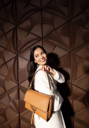 Tan leather handbag with decorative stitching, a gold hardware clasp, and a long strap, held by a person in a white coat against a geometric brown background.