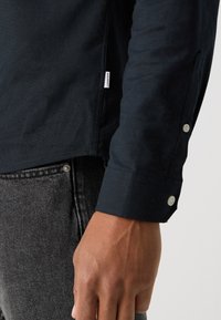 Camisa negra de manga larga con textura suave, puño abotonado y etiqueta visible. Combinada con vaqueros de mezclilla gris con un botón de metal.