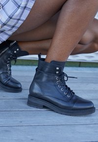 Botas negras de cuero hasta el tobillo con cordones, punta redondeada y un acabado texturizado. Presenta ojales plateados y un zipper lateral. Tacón bajo.