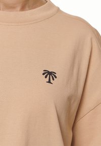 Světle hnědé bavlněné tričko s krátkým rukávem a kulatým výstřihem, s malou černou grafikou palmy na hrudi.