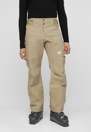 Pantaloni beige da esterno con tasche con cerniera sulle cosce, indossati con scarponi da sci neri e una maglia nera a maniche lunghe, visibile dalla vita in giù.