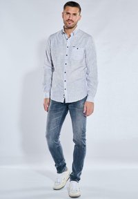 Hemd mit Knopfverschluss in hellgrau mit vertikalen Streifen, langen Ärmeln und einer Brusttasche, kombiniert mit blauen Jeans und weißen Turnschuhen.