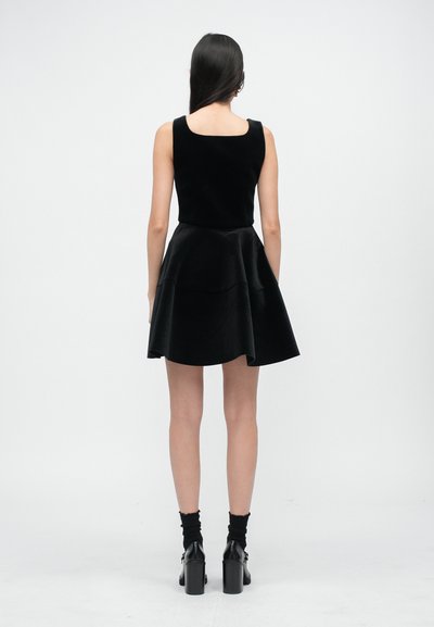 Femme aux longs cheveux foncés portant une robe noire sans manches, des chaussettes noires et des chaussures noires à talons, debout de dos devant un fond uni.