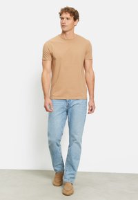 Beige T-Shirt mit kurzen Ärmeln in taillierter Form, kombiniert mit hellblauen Jeans und braunen Slippern, stehend auf einem neutralen Boden.