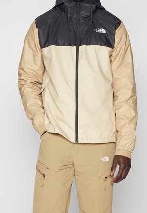 Mężczyzna w beżowej i czarnej kurtce z kapturem marki North Face oraz beżowych spodniach typu cargo North Face z zasuwanymi kieszeniami, stojący z jedną ręką w kieszeni.