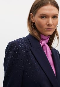Blazer della marina con dettagli decorati sulle spalle, caratterizzato da sfumature di pietre blu. Abbinato a una blusa rosa con collo a cravatta.