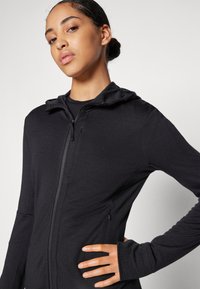 Melnā kapucēta sporta jaka ar pieguļošu dizainu, mīkstu tekstūru un kapuci. Iekļauj divpusēju rāvējslēdzēju un sānu kabatas, ar garām piedurknēm un caurumiem īkšķiem.