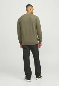 Pulover verde măsliniu cu guler rotund, cu manșete și tiv cu nervuri, purtat cu pantaloni negri și adidași. Material moale, texturat.