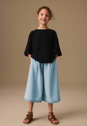 Young girl stands smiling with hands in pockets, wearing black top, light blue wide-leg jeans, and brown strappy sandals.