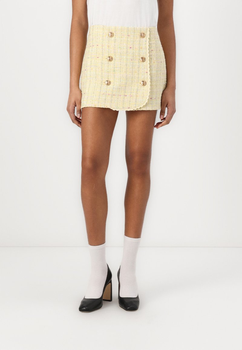 Gonna mini in tweed giallo con una flap laterale, bottoni dorati doppi e orlo scallop. Abbinata a calze bianche e tacchi neri.