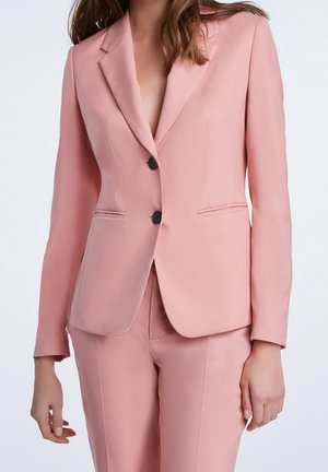 Femme portant un blazer rose clair cintré avec deux boutons noirs et un pantalon assorti et ajusté, debout sur un fond uni.