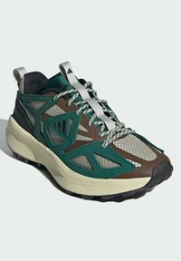 Scarpa sportiva verde e marrone con tomaia in rete, accentuata da lacci argentati, pannellatura texturizzata e una suola intermedia spessa di colore chiaro.