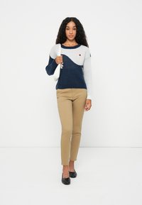 Strickpullover in Marineblau und Grau mit Wellenmuster, kombiniert mit khakifarbenen Hosen. Das Modell hält eine weiße Handtasche; das Schuhwerk hat eine Schleifchen-Details.