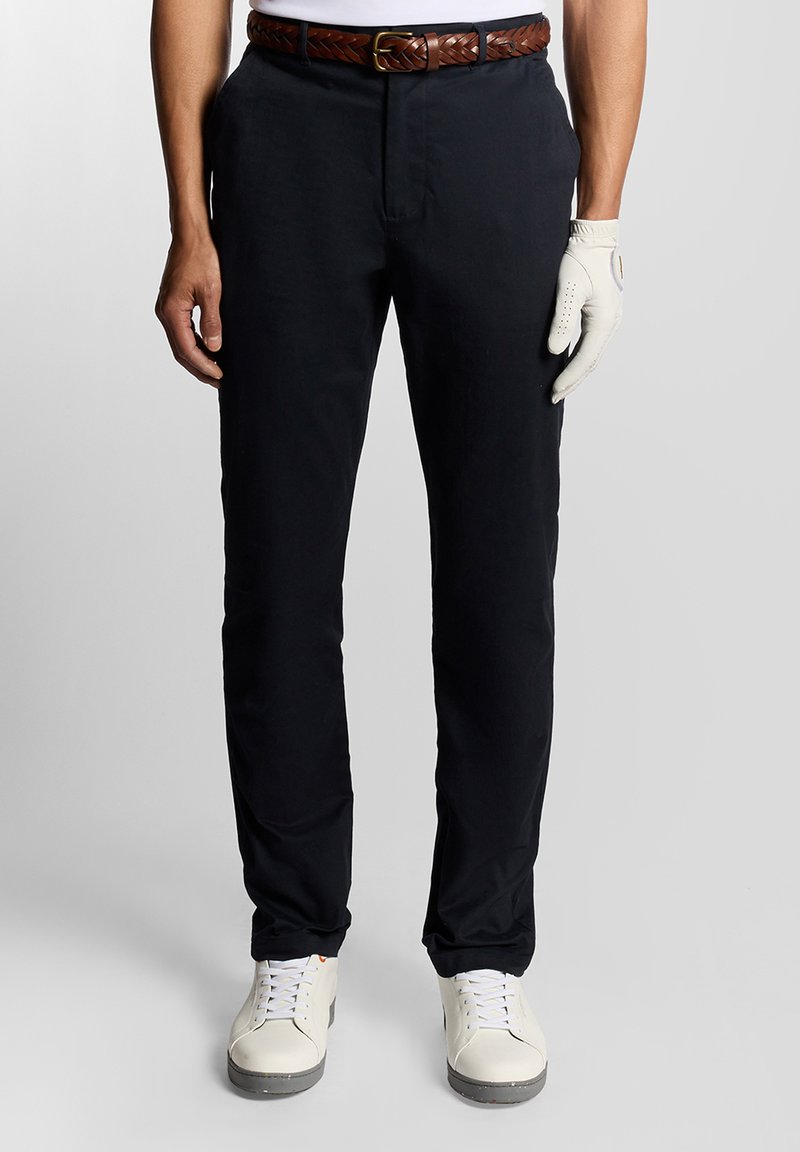 Schwarze Hose mit flachem Bund und Seitentaschen, kombiniert mit einem geflochtenen braunen Leder-Gürtel. Getragen mit einem weißen Hemd und Handschuhen.